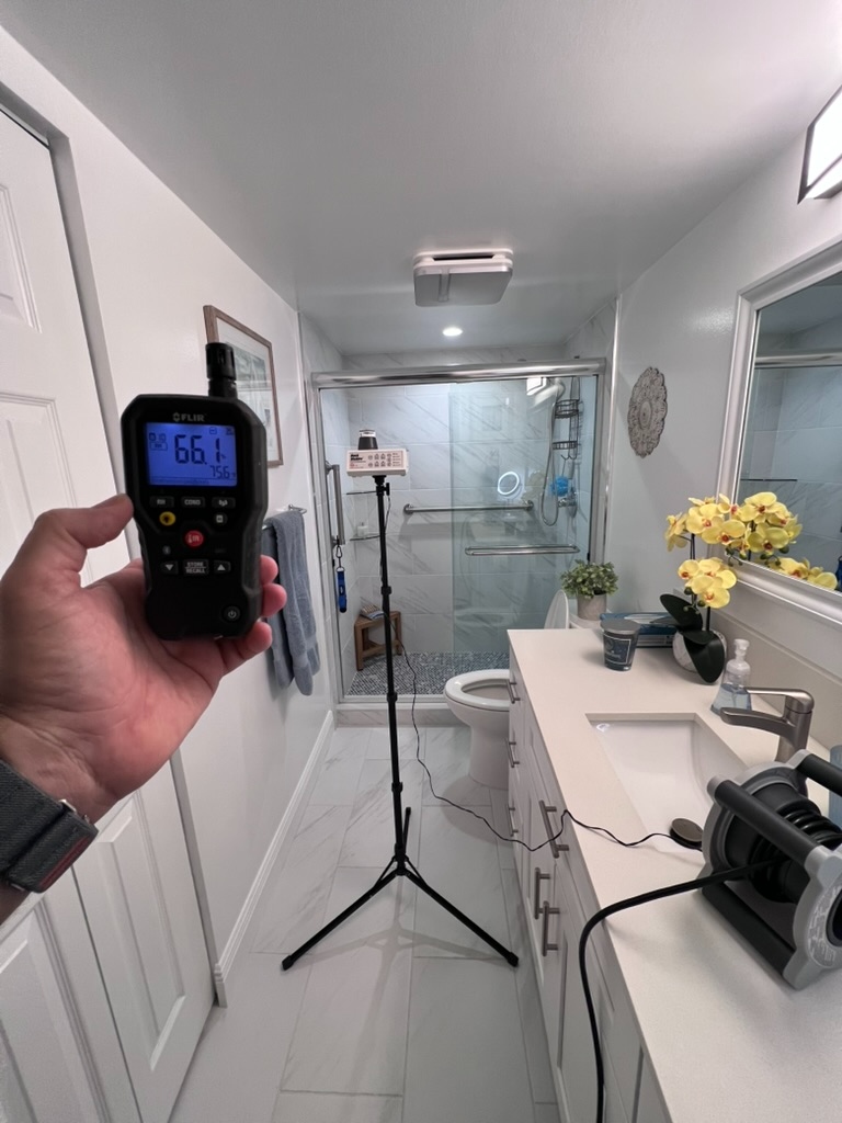 FLIR humidity meter showing 66.1 percent RH held in front of a bathroom setup with air sampling equipment on a tripod