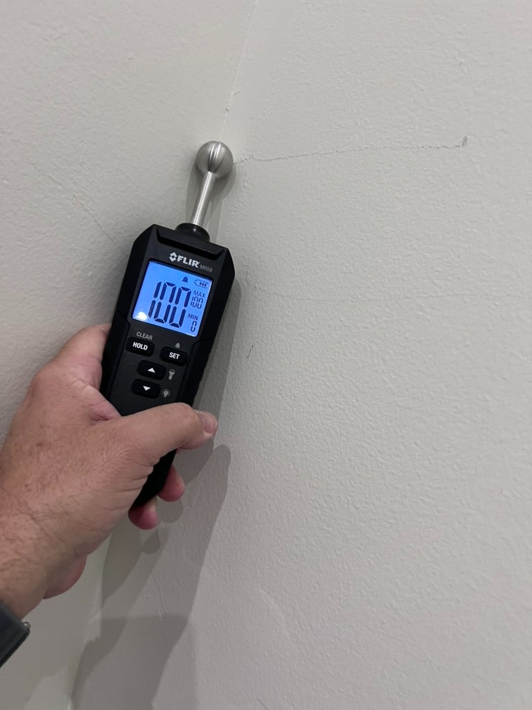 FLIR MR59 moisture meter pressed against a cracked wall showing a reading of 100 indicating saturated material