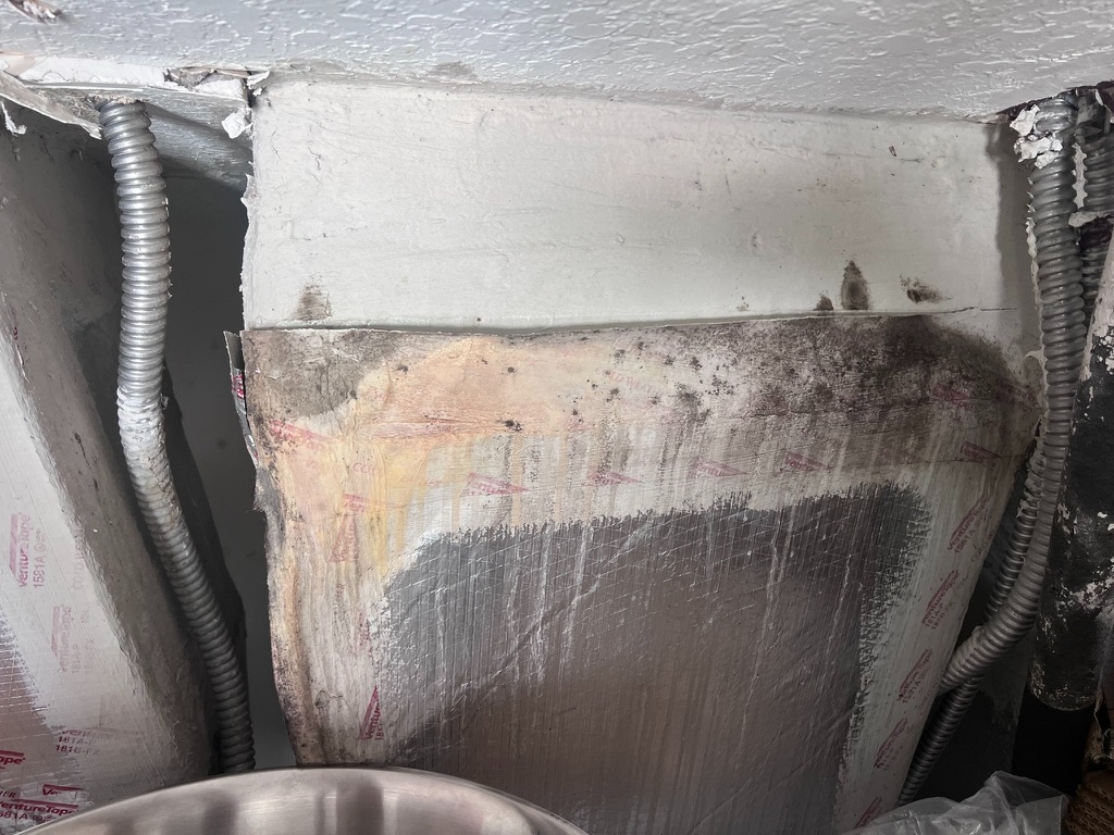 Wide view of the HVAC plenum insulation panel showing dark mold growth concentrated along the top edge where the panel meets the ceiling framing, with visible tape seams