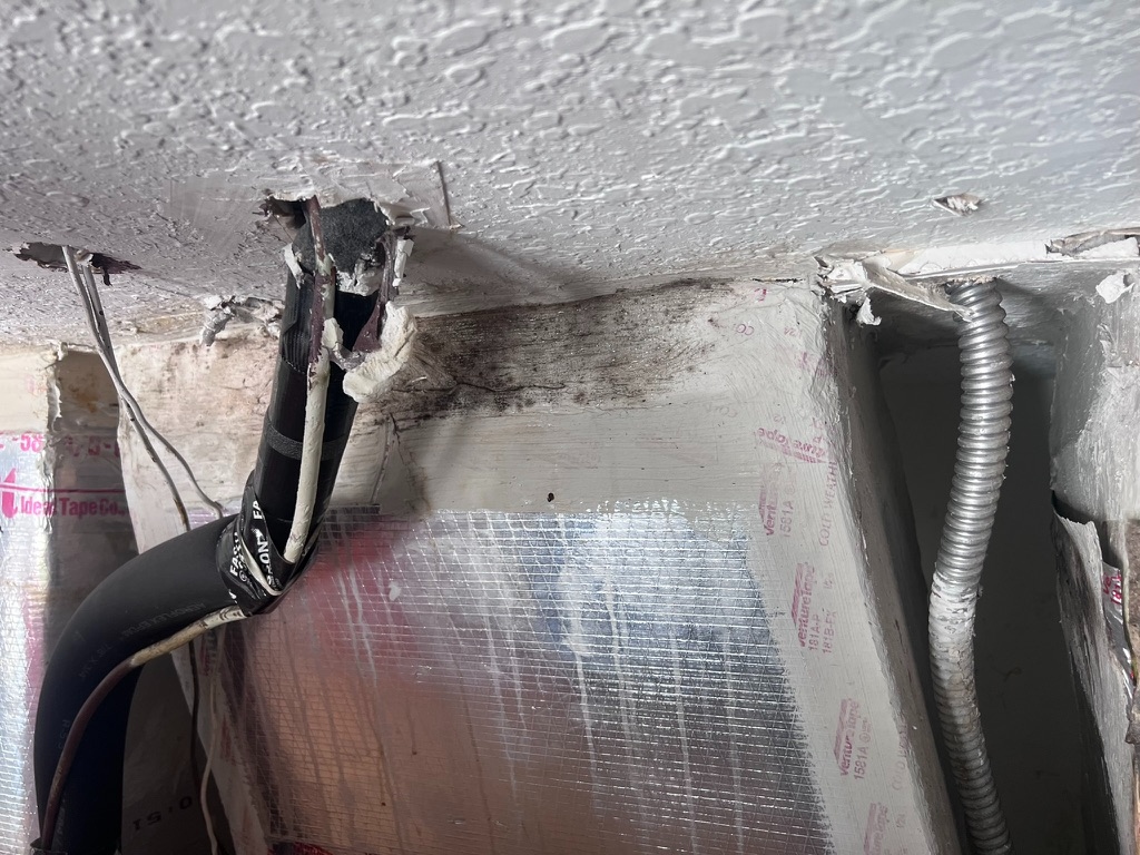Close-up of black HVAC tape wrapped around a duct connection at a ceiling penetration, with dark mold staining visible on the insulation panel below and beside it