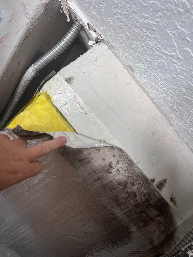 A hand pointing directly at a section of failed and separated tape seal at a duct connection, with mold visible below the gap and yellow fiberglass insulation exposed where the tape has pulled away